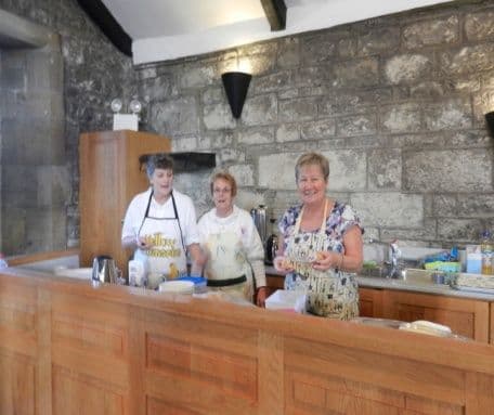 Three women in aprons serving soup and drinks from a church kitchen counter