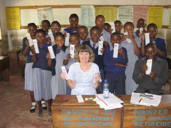 Pupils in a classroom holding up handmade bookmarks around a visiting woman seated at a donated desk