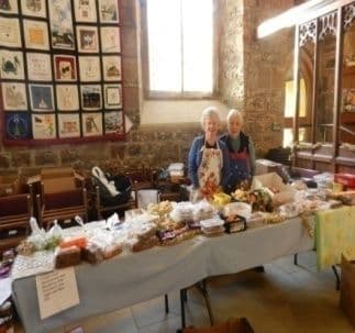 Two women behind a long table of cakes and baking in a church hall
