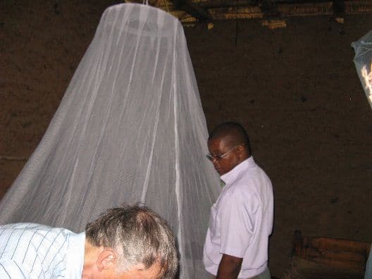 Two men hanging a white mosquito net over a bed inside a home