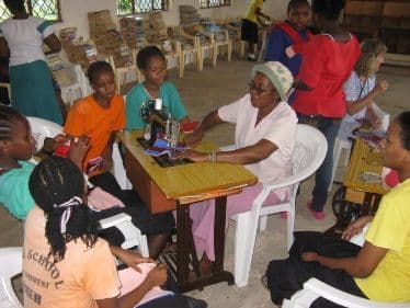 Woman at a sewing machine with girls gathered around her in the library