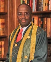 Man in green judicial robes with gold trim and a red tie, in front of bookshelves