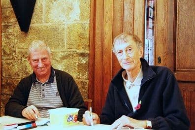 Two older men seated at a table in a church hall, one writing on a sheet