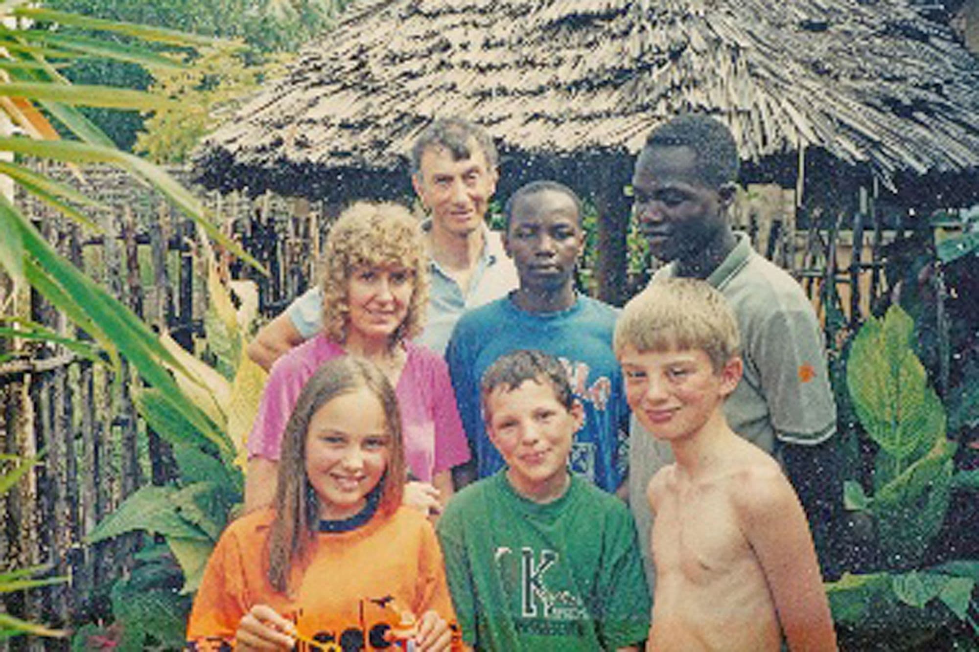 A woman, three children and two young Kenyan men posing in front of a thatched hut
