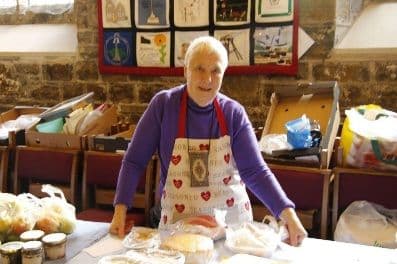 Woman in a heart-print apron behind a table of cakes and jars at a church sale