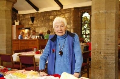 Older woman in a blue cardigan standing at a stall in a stone church hall