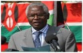 Grey-haired man in a grey suit speaking into microphones in front of a Kenyan flag