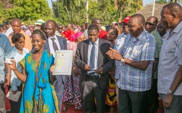 A woman in a blue dress holds up a certificate beside men in suits amid a crowd outdoors