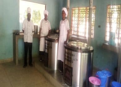 Three cooks in white uniforms and hats standing beside large steel cooking pots in the school kitchen