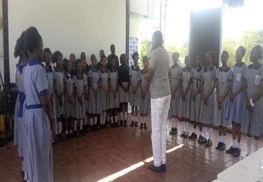 Pupils in school uniform standing in rows singing, led by a man in a white shirt