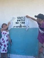 A woman and a man pull a white cloth off a painted plaque on a classroom wall to open the building