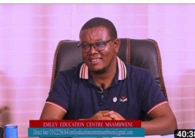 Man in a navy polo shirt seated in a TV studio with an Emley Education Centre Msambweni banner on screen