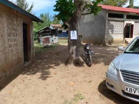 An eye chart fixed to a tree trunk in a dirt yard between buildings, with a parked motorbike