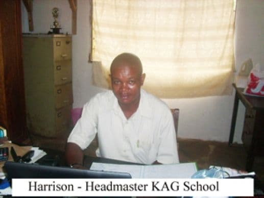 The headmaster seated at his office desk with a filing cabinet and trophy behind him