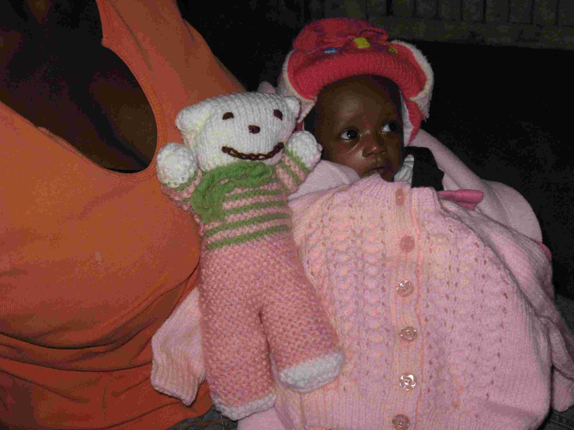A baby in a knitted pink hat and cardigan beside a knitted teddy bear