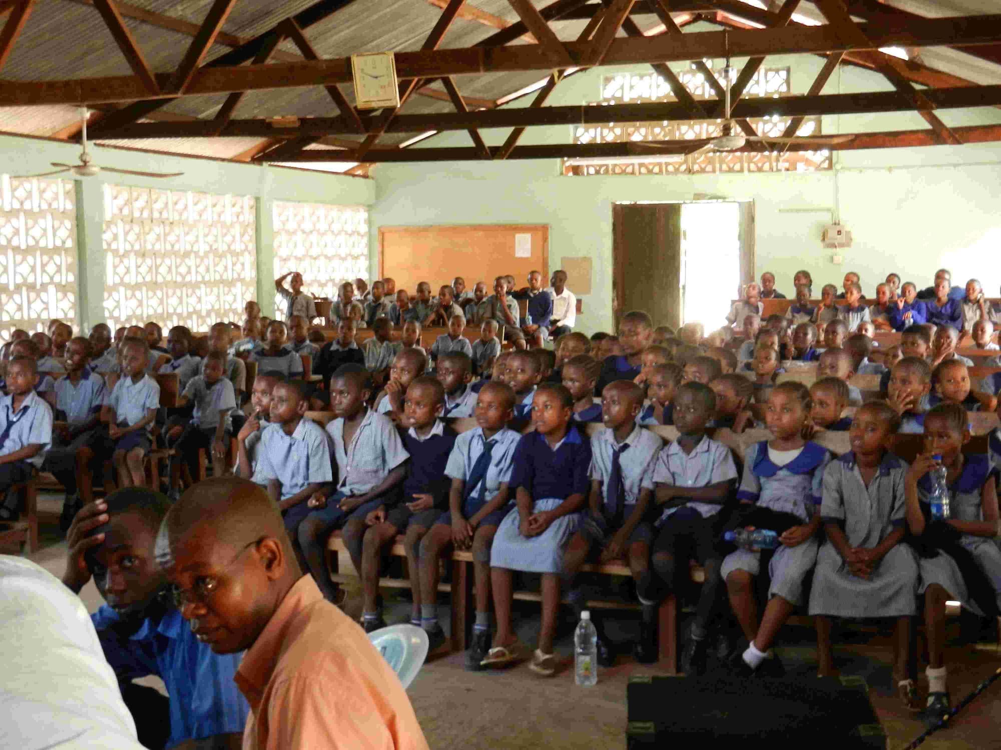A hall packed with pupils in uniform watching a programme