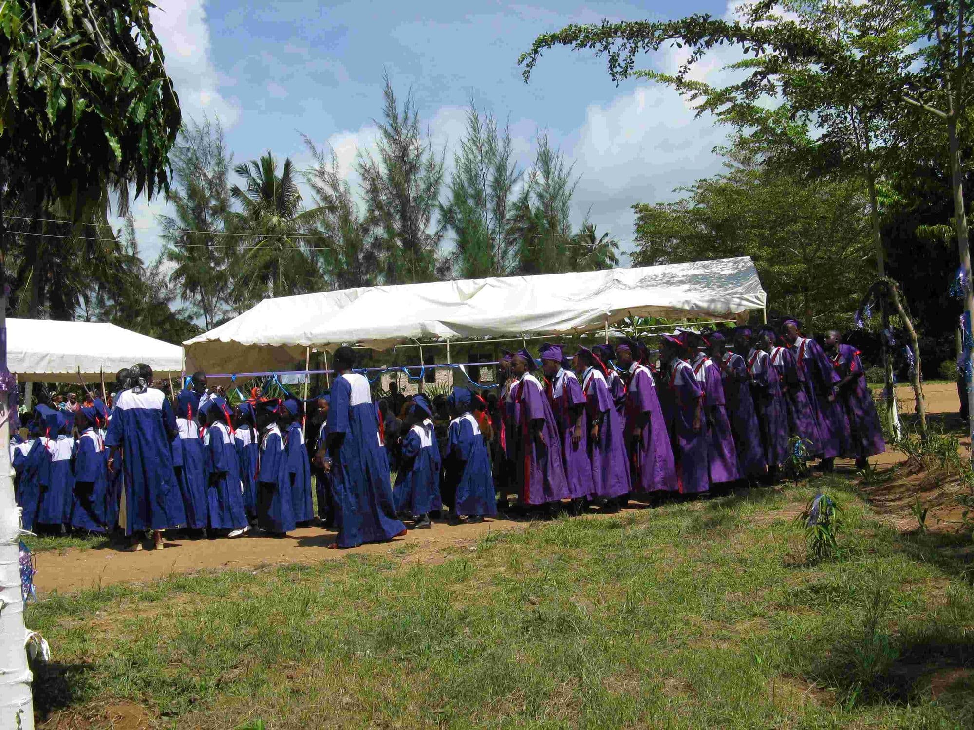 Graduates in blue and purple gowns lined up beside white marquees on the school grounds