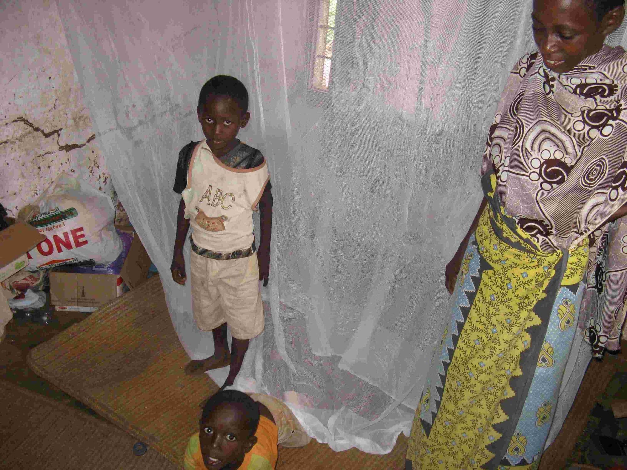 A boy beside a mosquito net hung over a mattress on the floor of his home