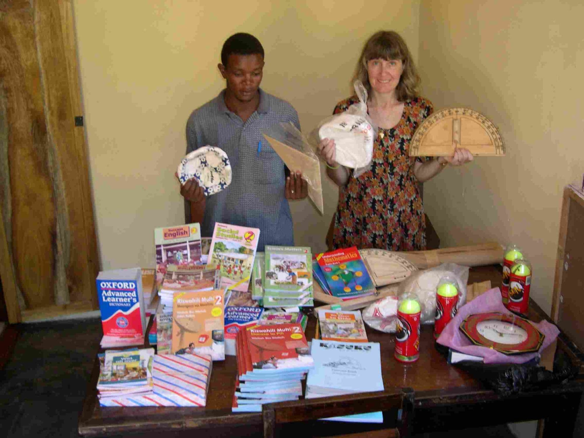 Two adults holding up donated books, school supplies and maths equipment laid out on a table