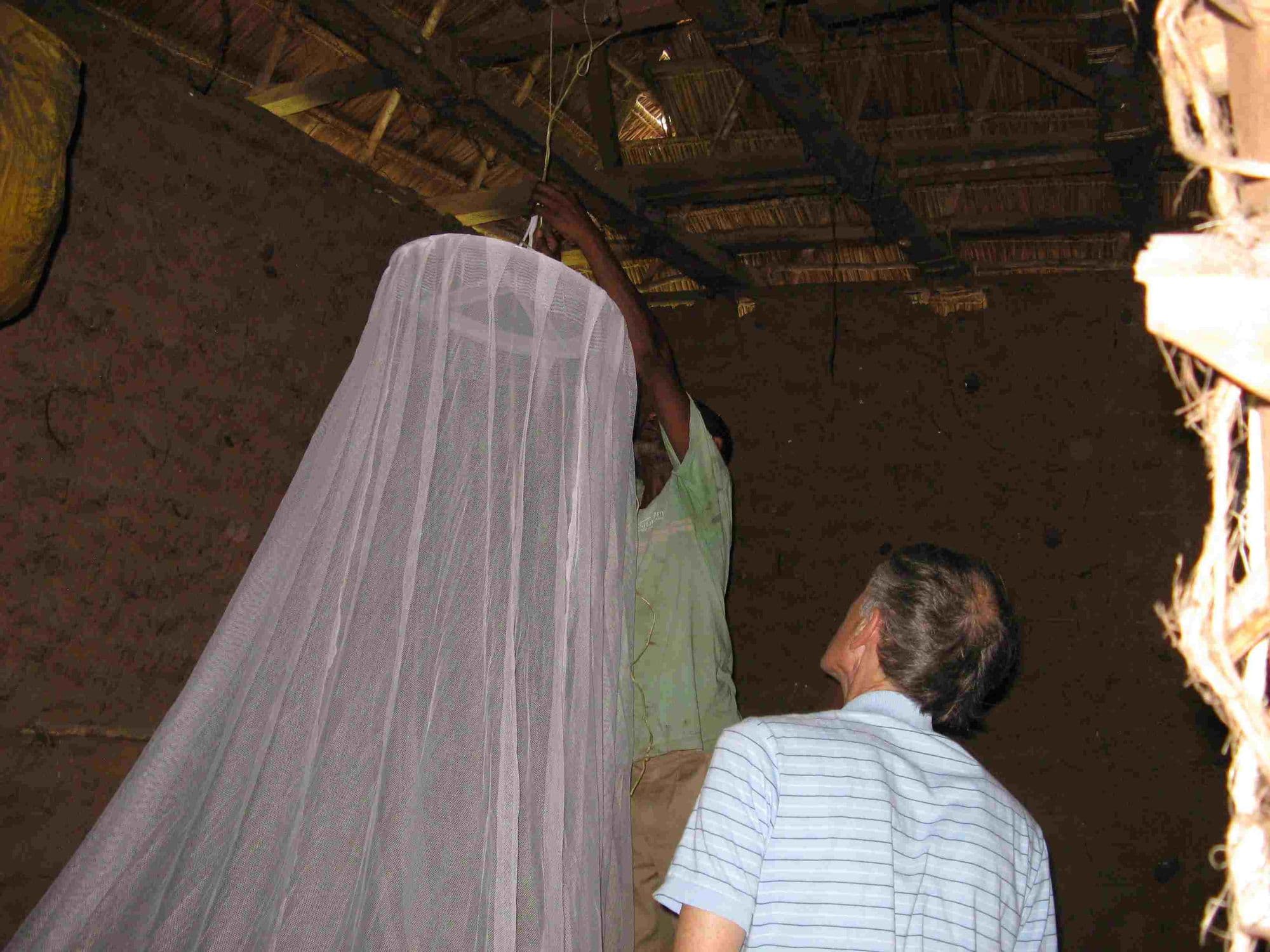 A visitor helping hang a mosquito net from the roof of a mud house