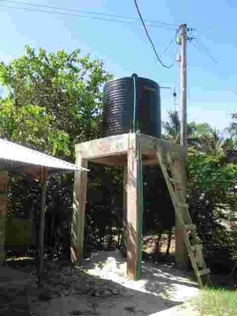 A black water tank raised on a wooden tower beside a school building
