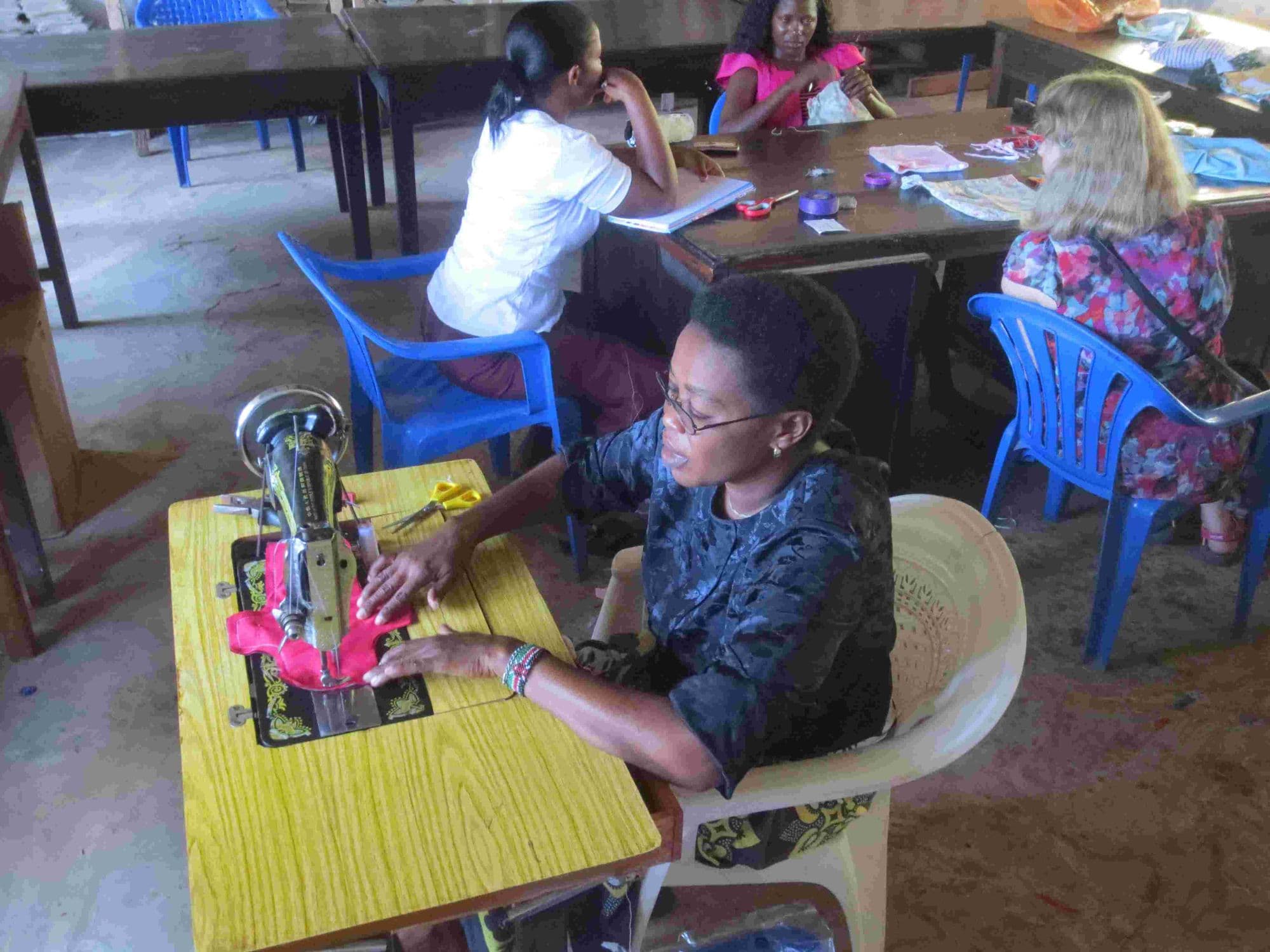 A woman sewing at a treadle machine while others work at tables behind her