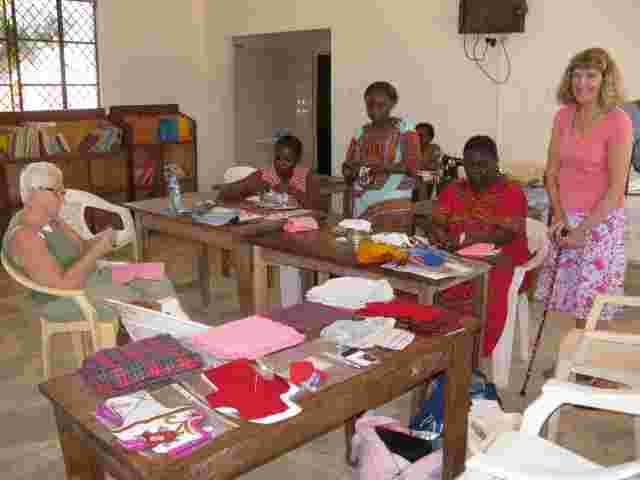 Women sewing sanitary wear at tables in a church hall with visitors helping