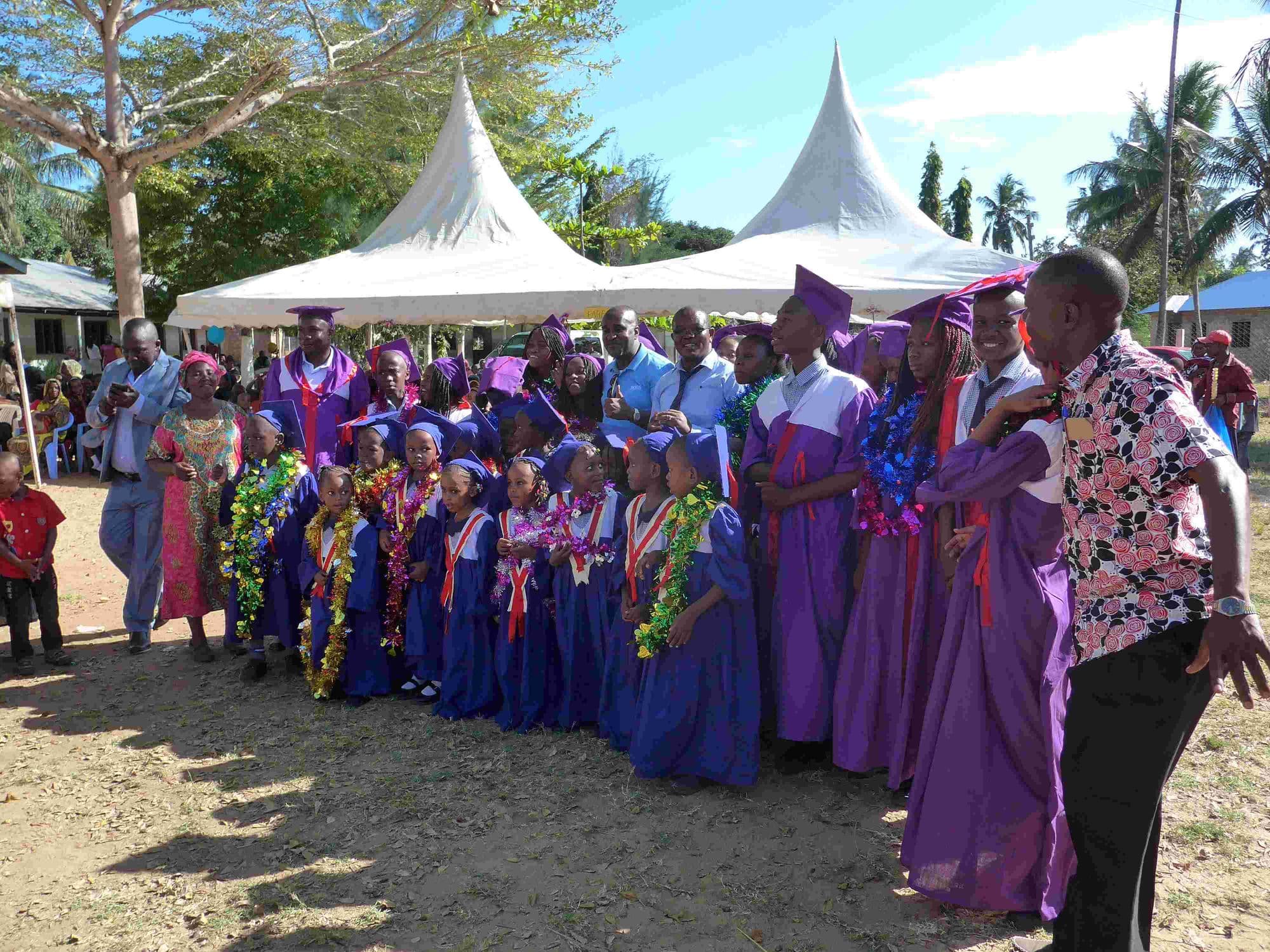 Graduates in blue and purple gowns with garlands lined up in front of white marquees