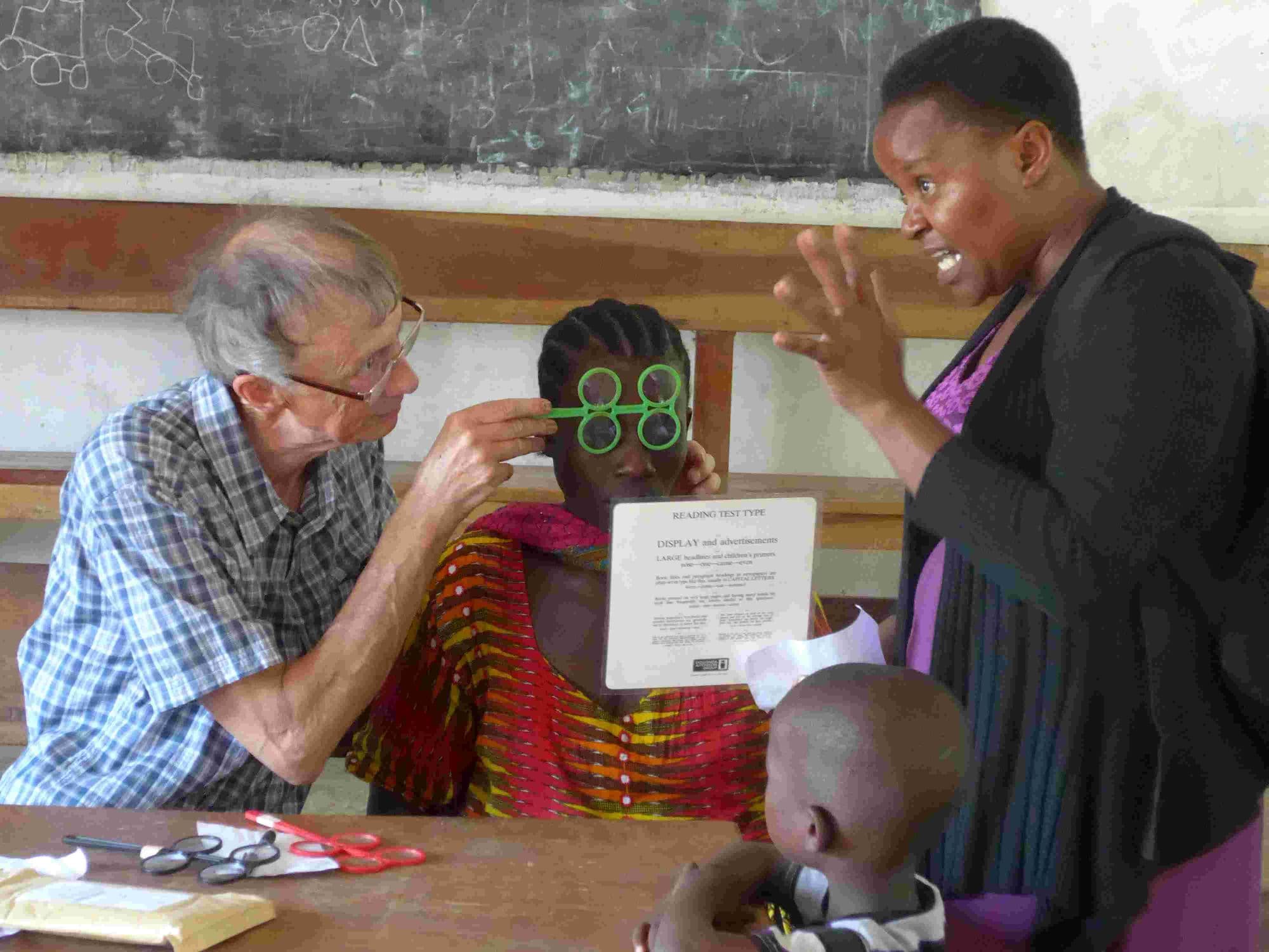 A visitor testing a woman's eyesight with trial lenses while a teacher helps
