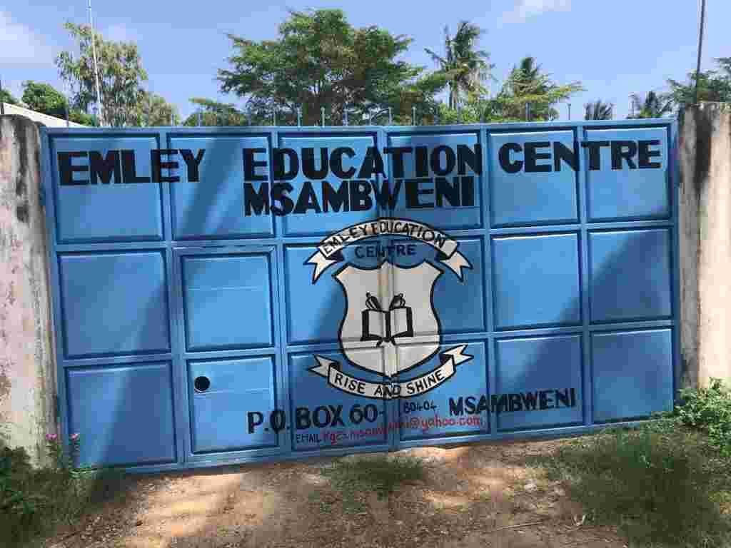 The blue school gate painted with the name Emley Education Centre Msambweni and the school crest