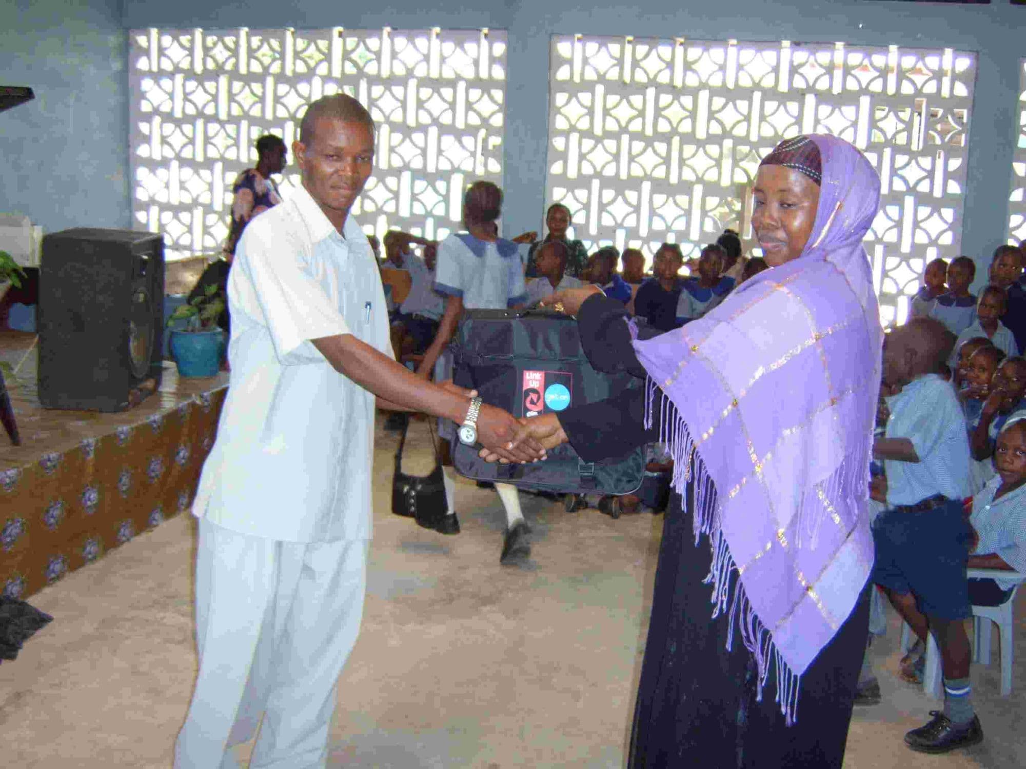 A man in white handing a gift to a woman in a lilac headscarf in front of seated pupils