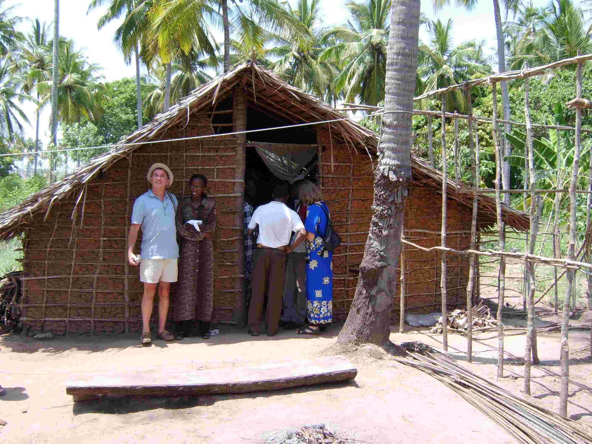 Visitors standing at the doorway of a thatched mud-walled house among palm trees