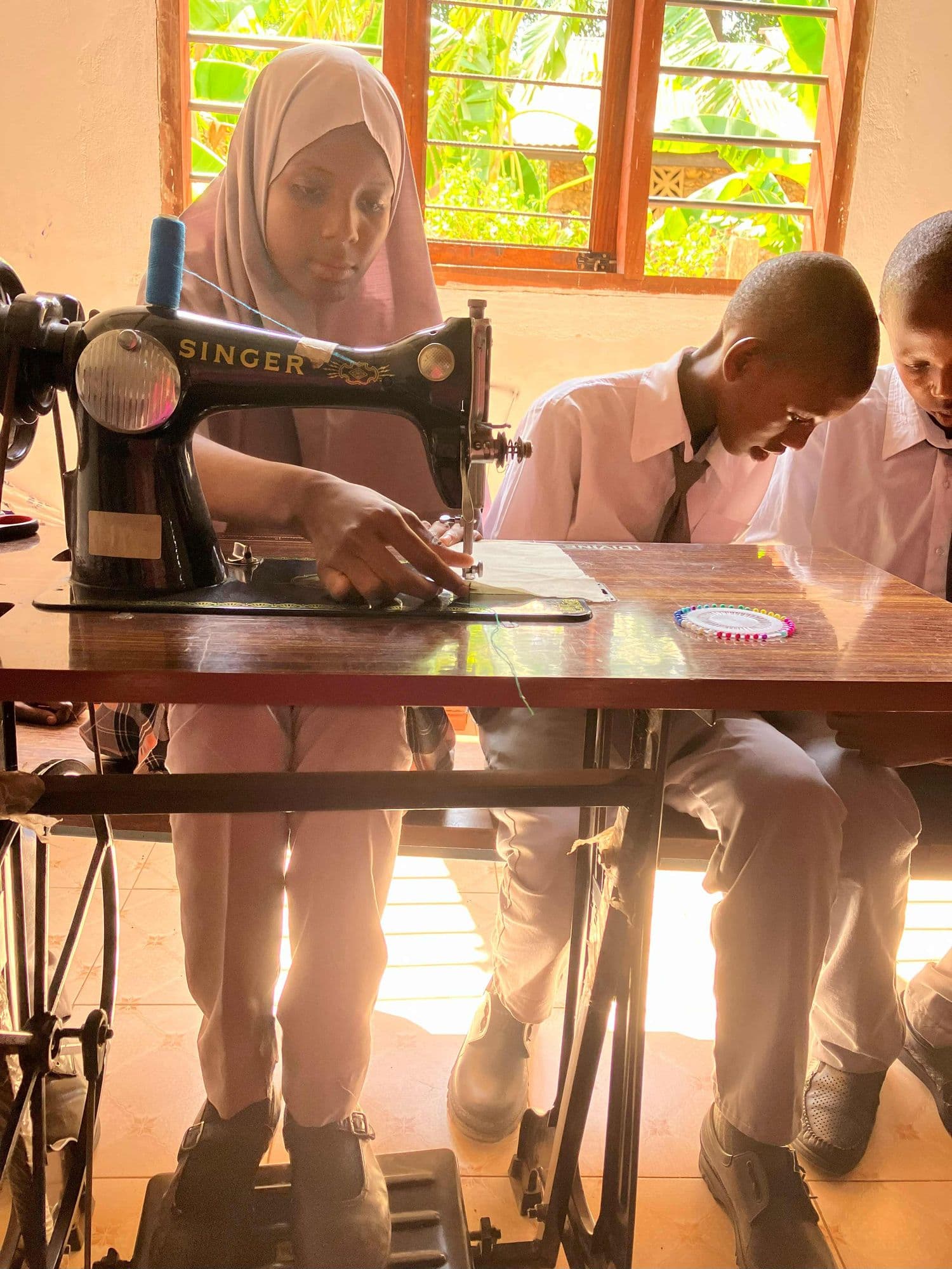 A schoolgirl sewing at a treadle sewing machine while a boy watches