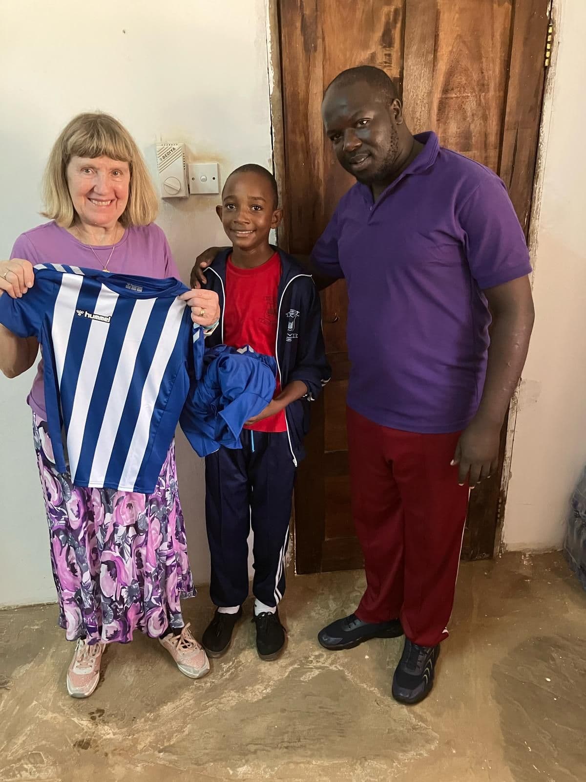 A visitor and a teacher with a boy holding up a blue and white striped football shirt