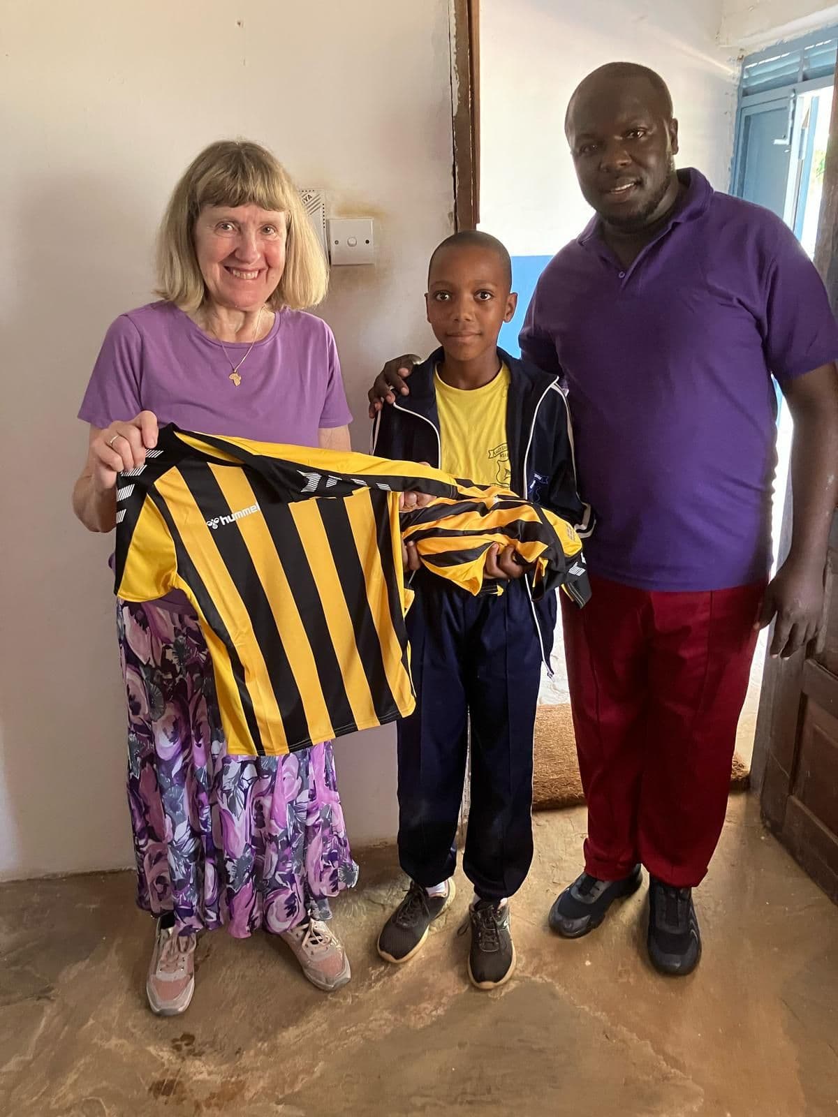 A visitor and a teacher with a boy holding up a black and yellow striped shirt