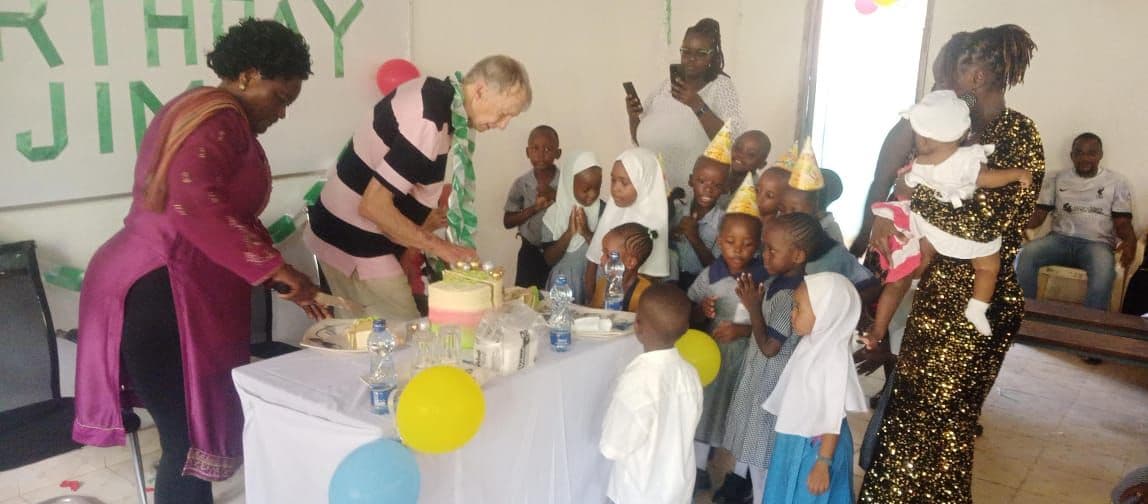 A visitor cutting the birthday cake surrounded by teachers and young pupils in party hats