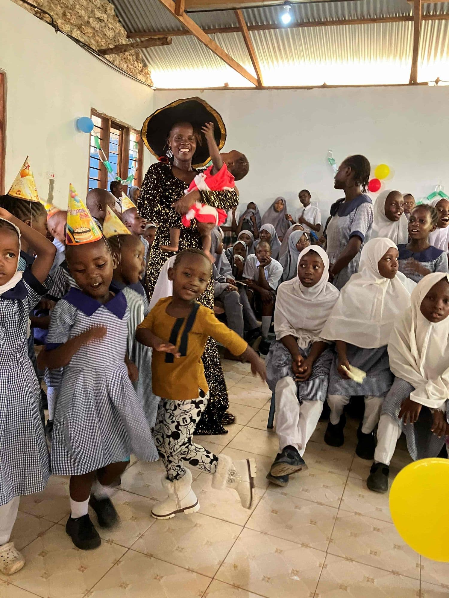 Young pupils in party hats dancing in front of the seated school