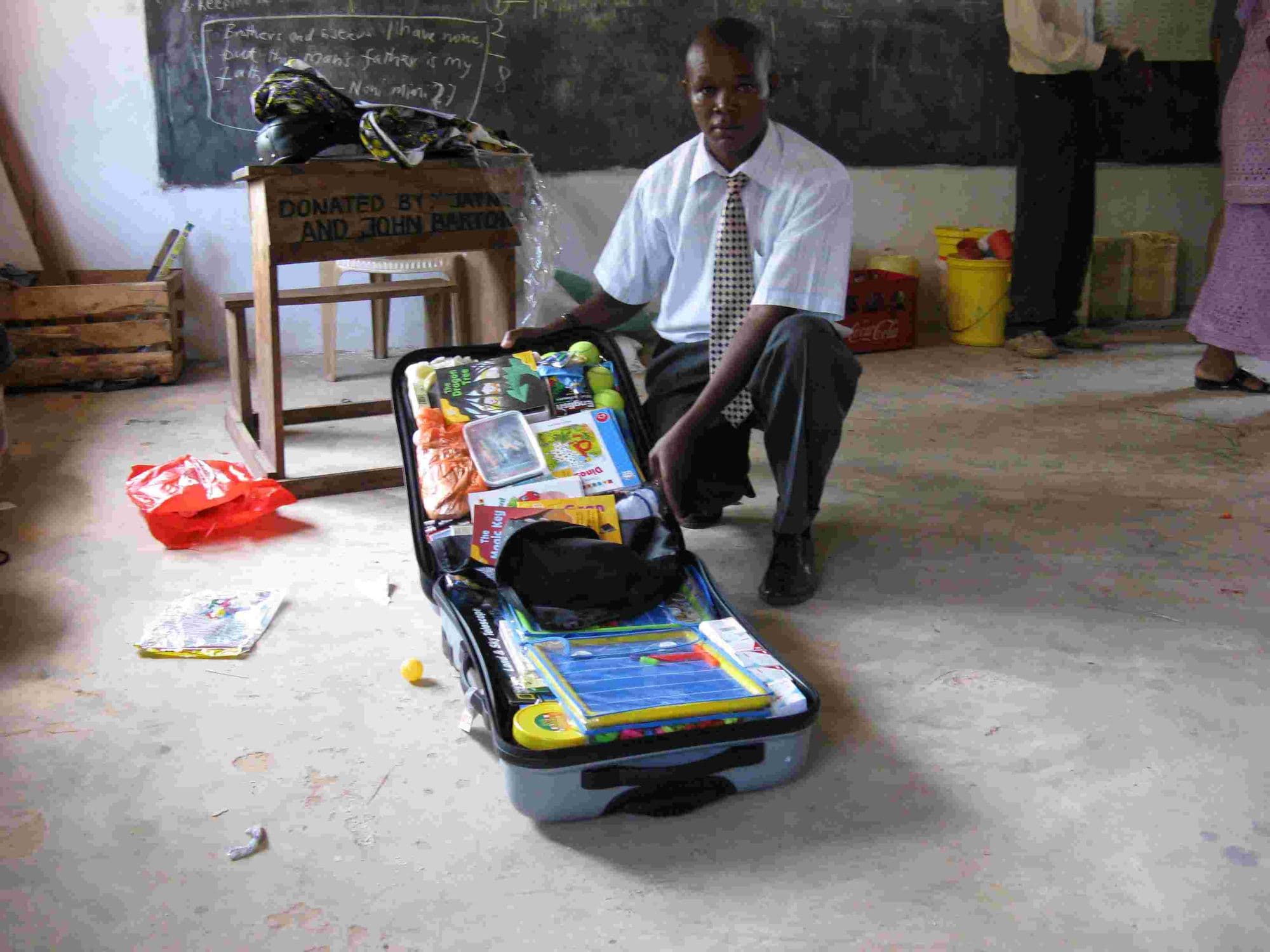 A man kneeling by an open suitcase packed with school supplies and toys on a classroom floor
