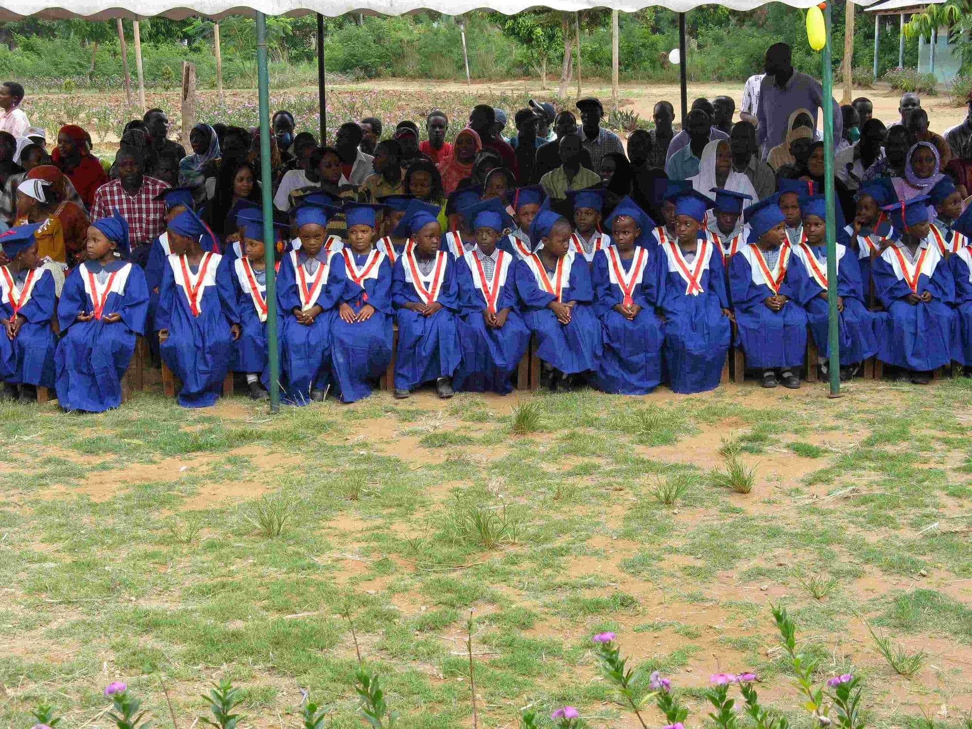 Rows of graduates in blue gowns seated under a canopy with parents behind