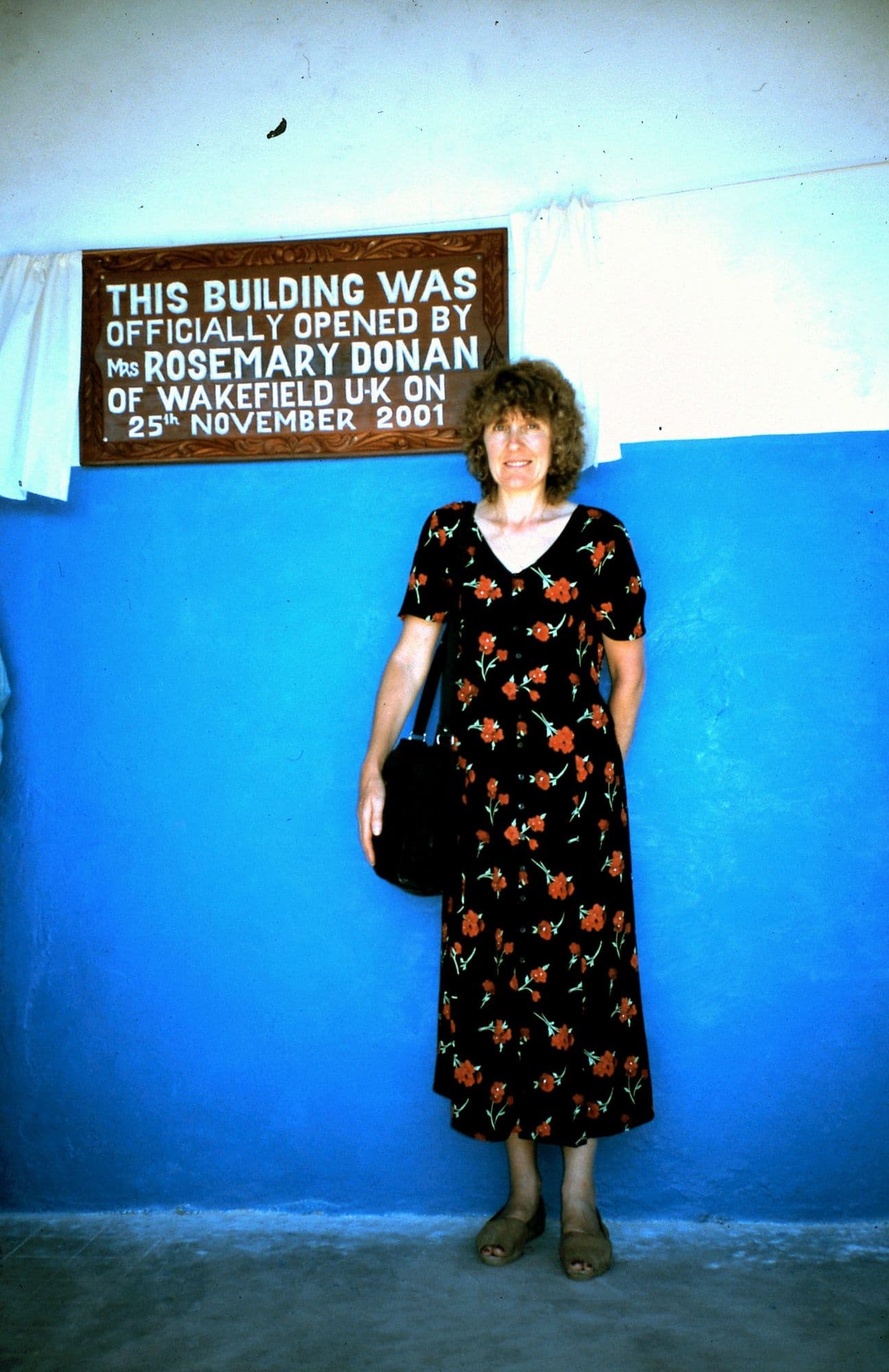 A woman in a floral dress standing below the plaque recording the nursery's opening on 25 November 2001