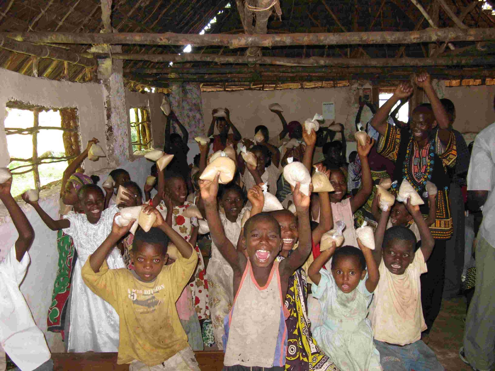Excited children holding up packets of food above their heads in a thatched hall