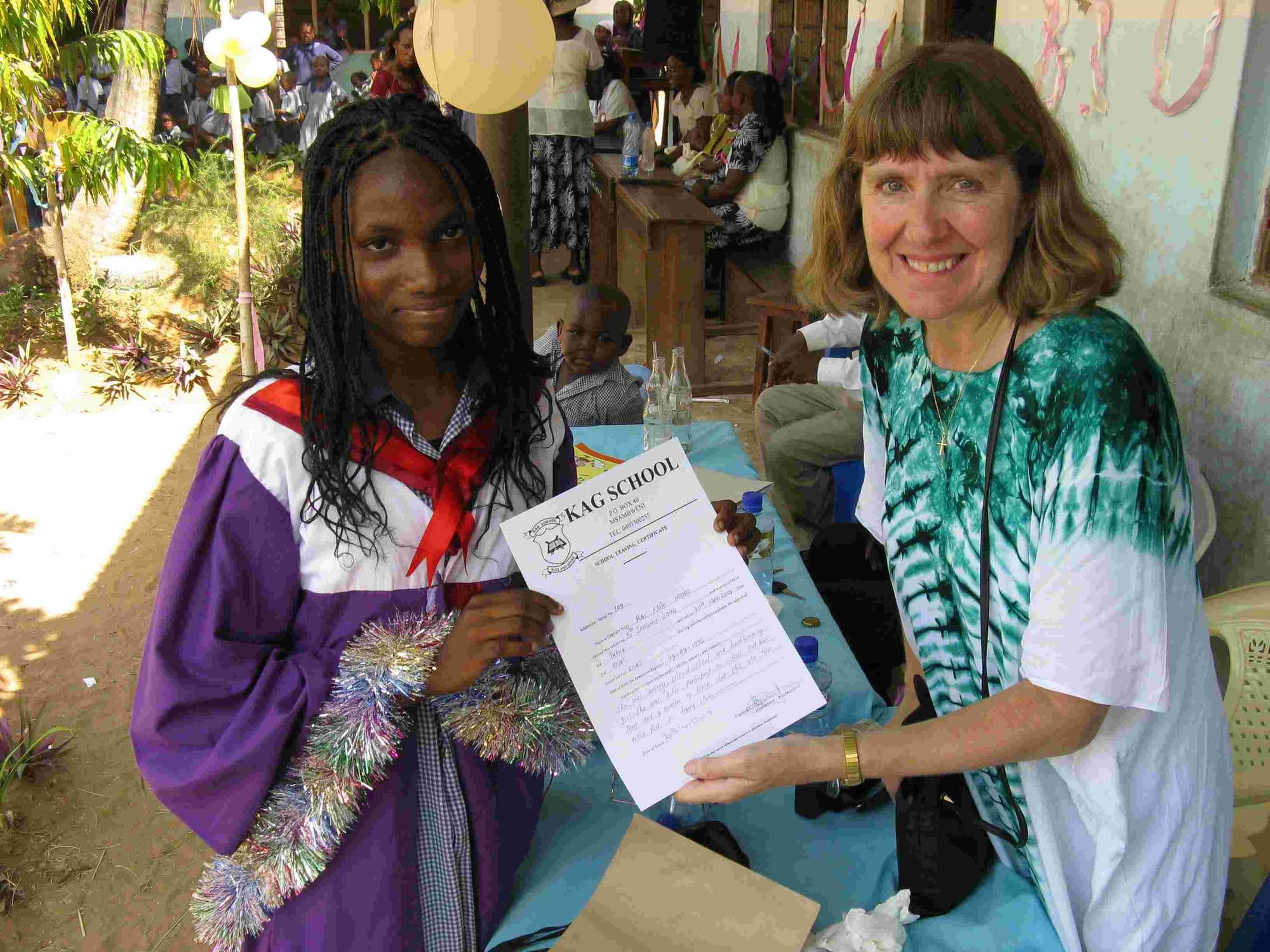 A girl in a purple graduation gown holding a certificate beside a smiling visitor