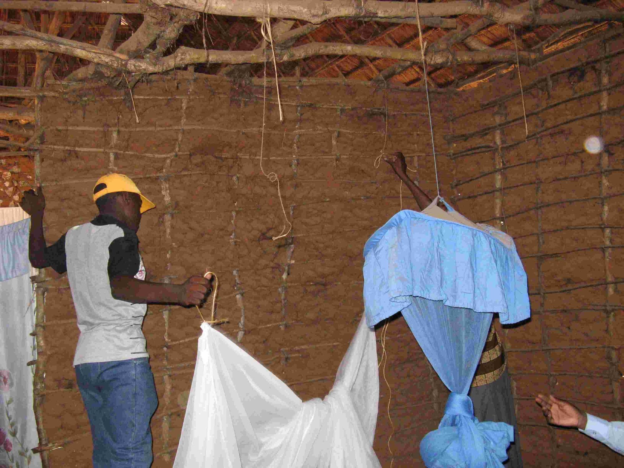 A man fixing a mosquito net to the roof poles inside a mud house
