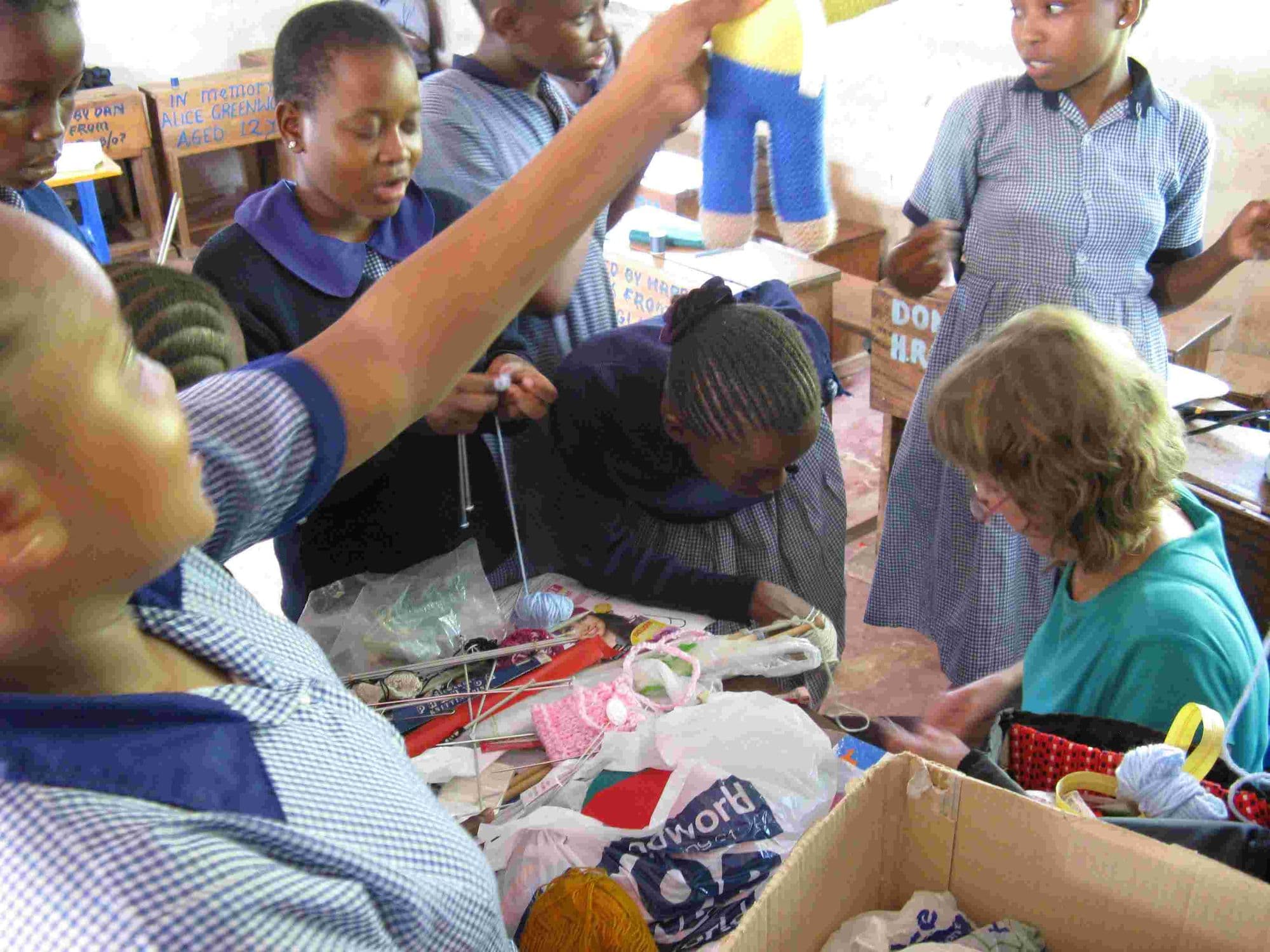 A visitor helping schoolgirls with knitting around a box of wool