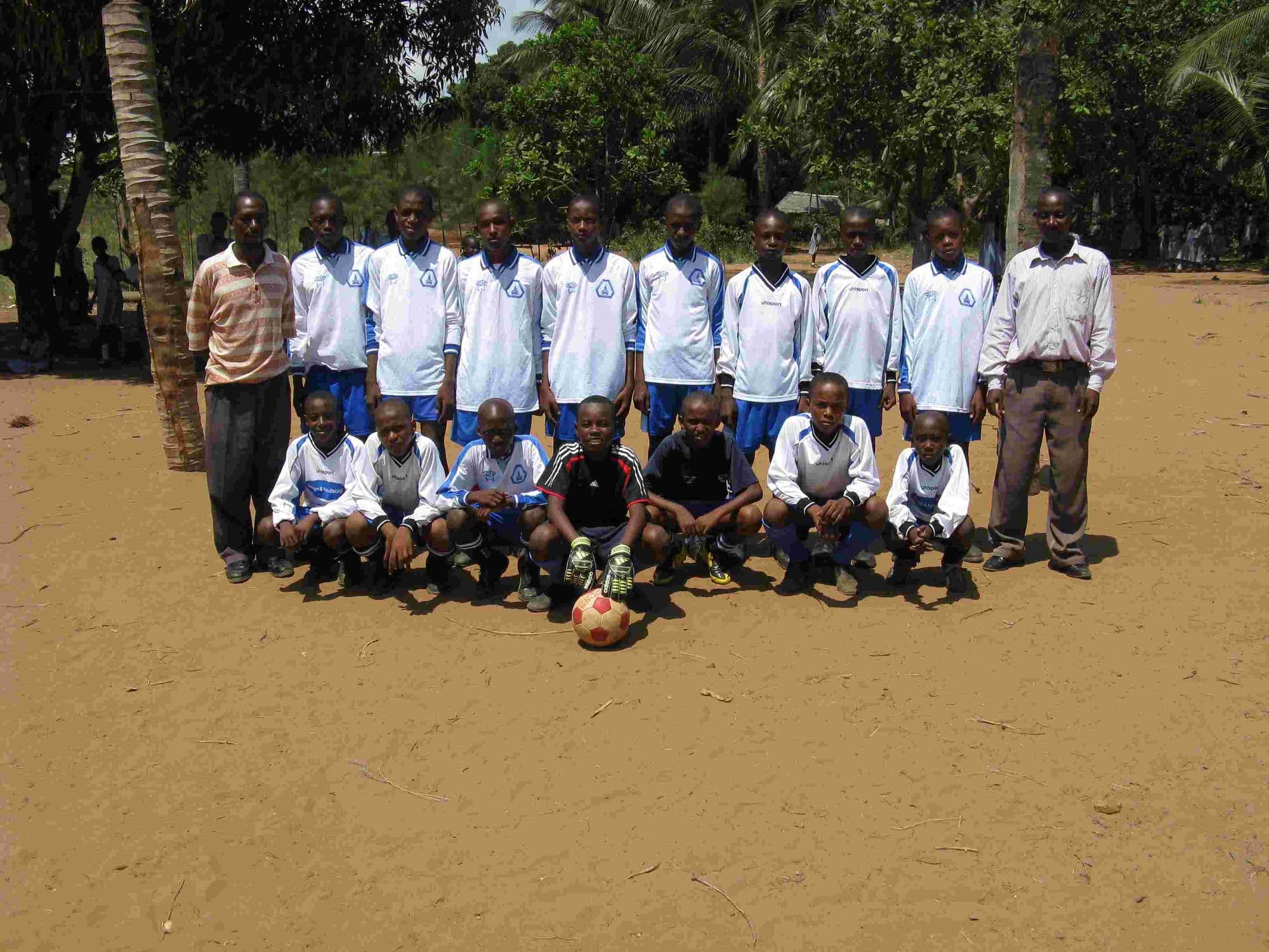 A football team in white kit lined up with two teachers on a sandy pitch