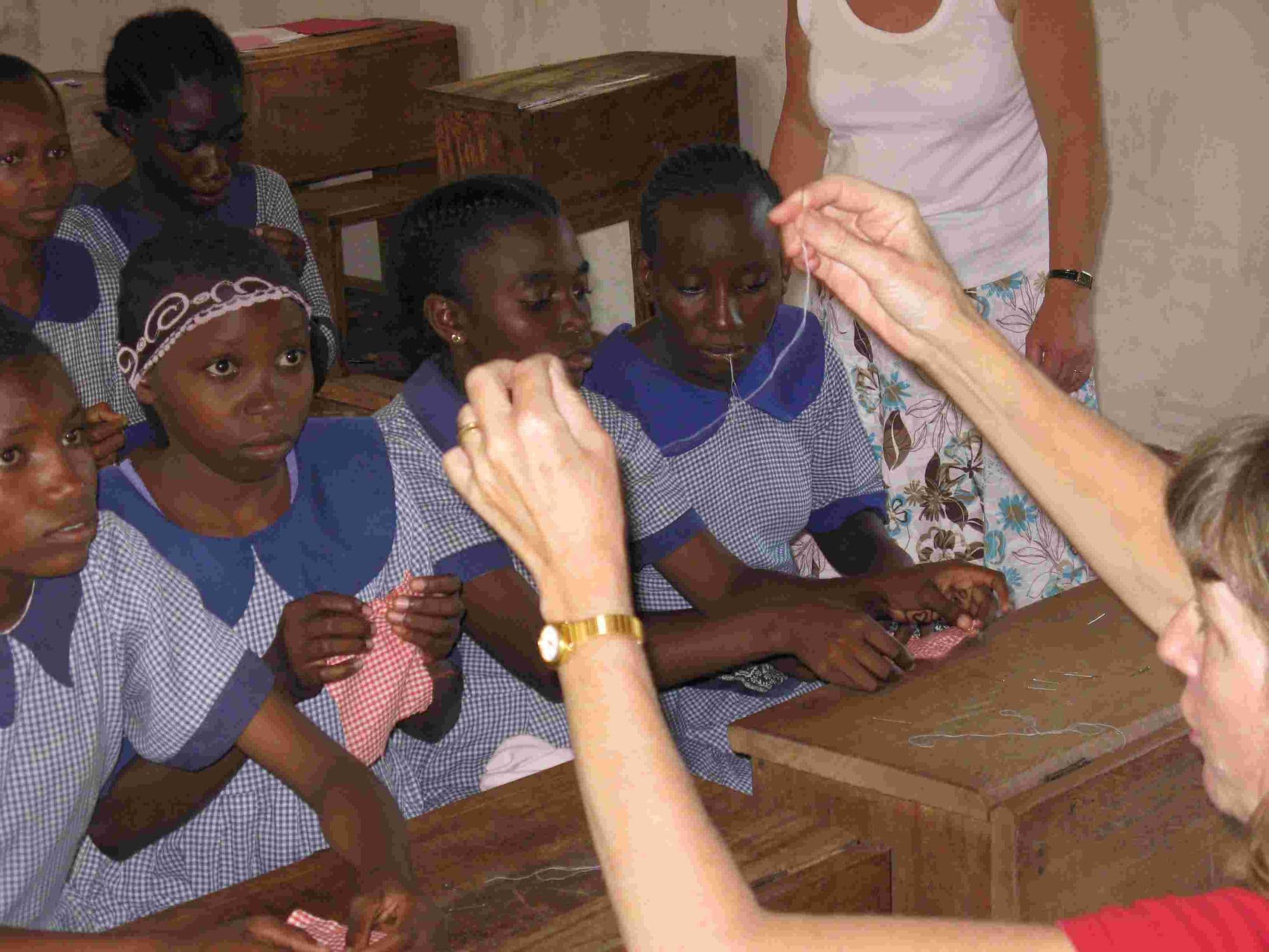 Schoolgirls threading needles at their desks while a visitor's hand demonstrates