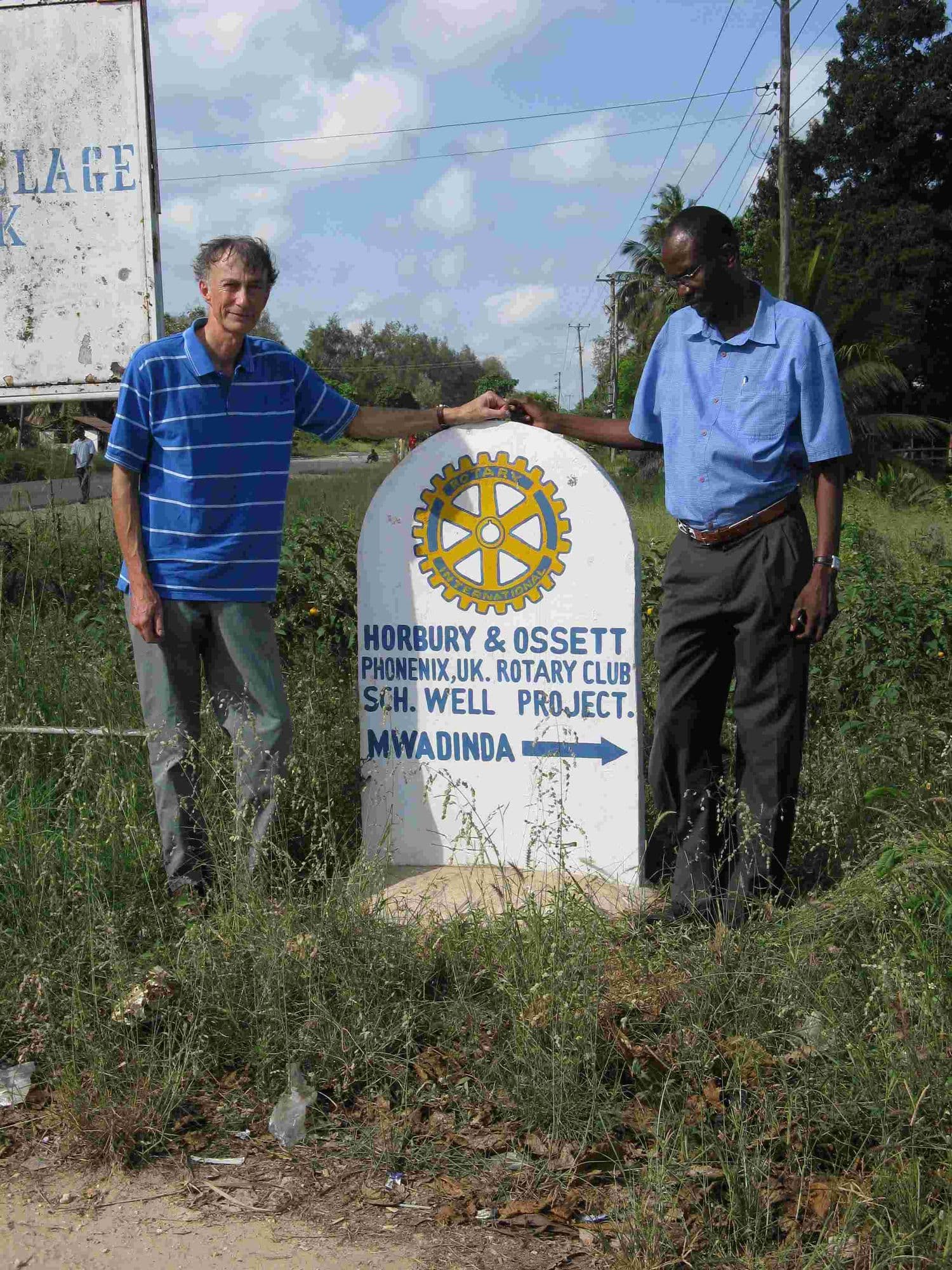 Two men beside a roadside marker for the Horbury & Ossett Phoenix Rotary Club well project, Mwadinba