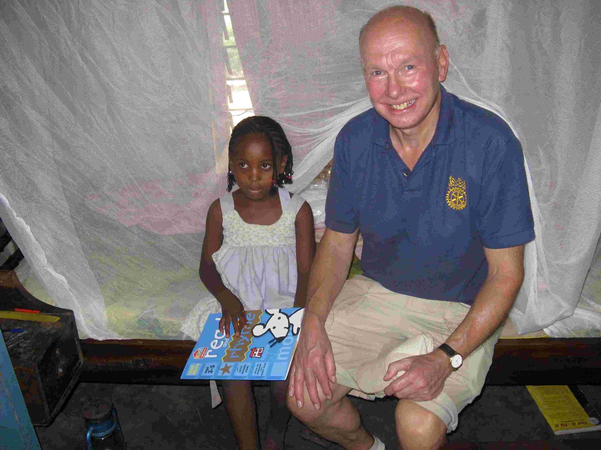 A visitor sitting with a young girl under a mosquito net, holding a picture book