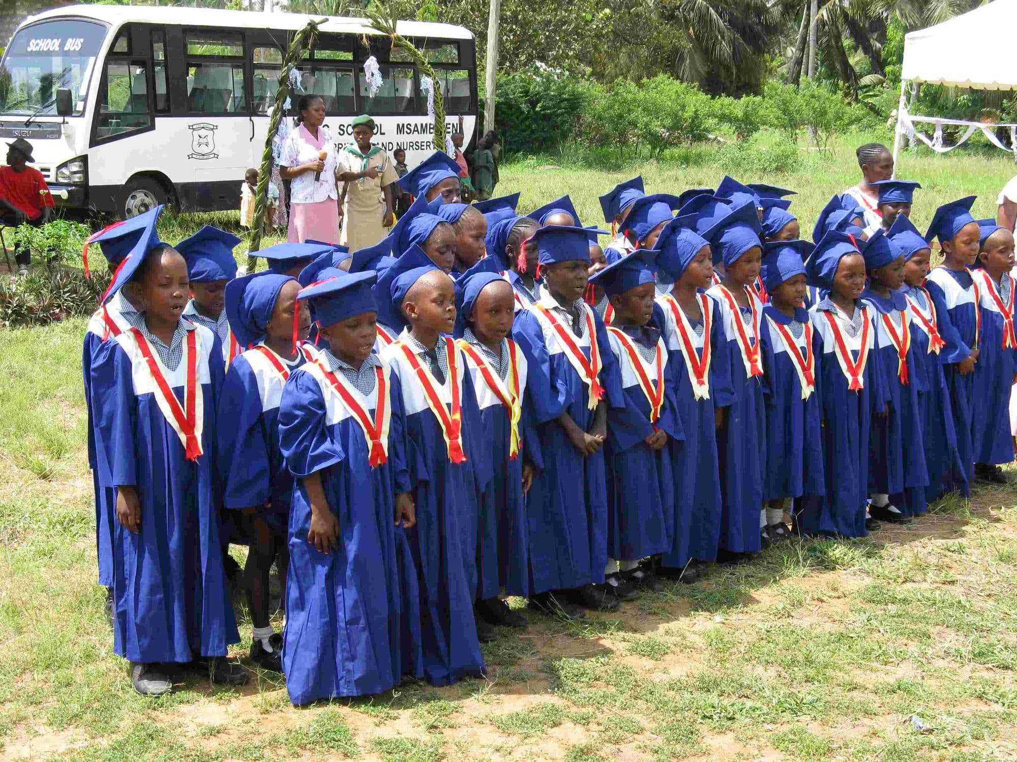 Nursery graduates in blue gowns and mortarboards lined up outside beside a school bus