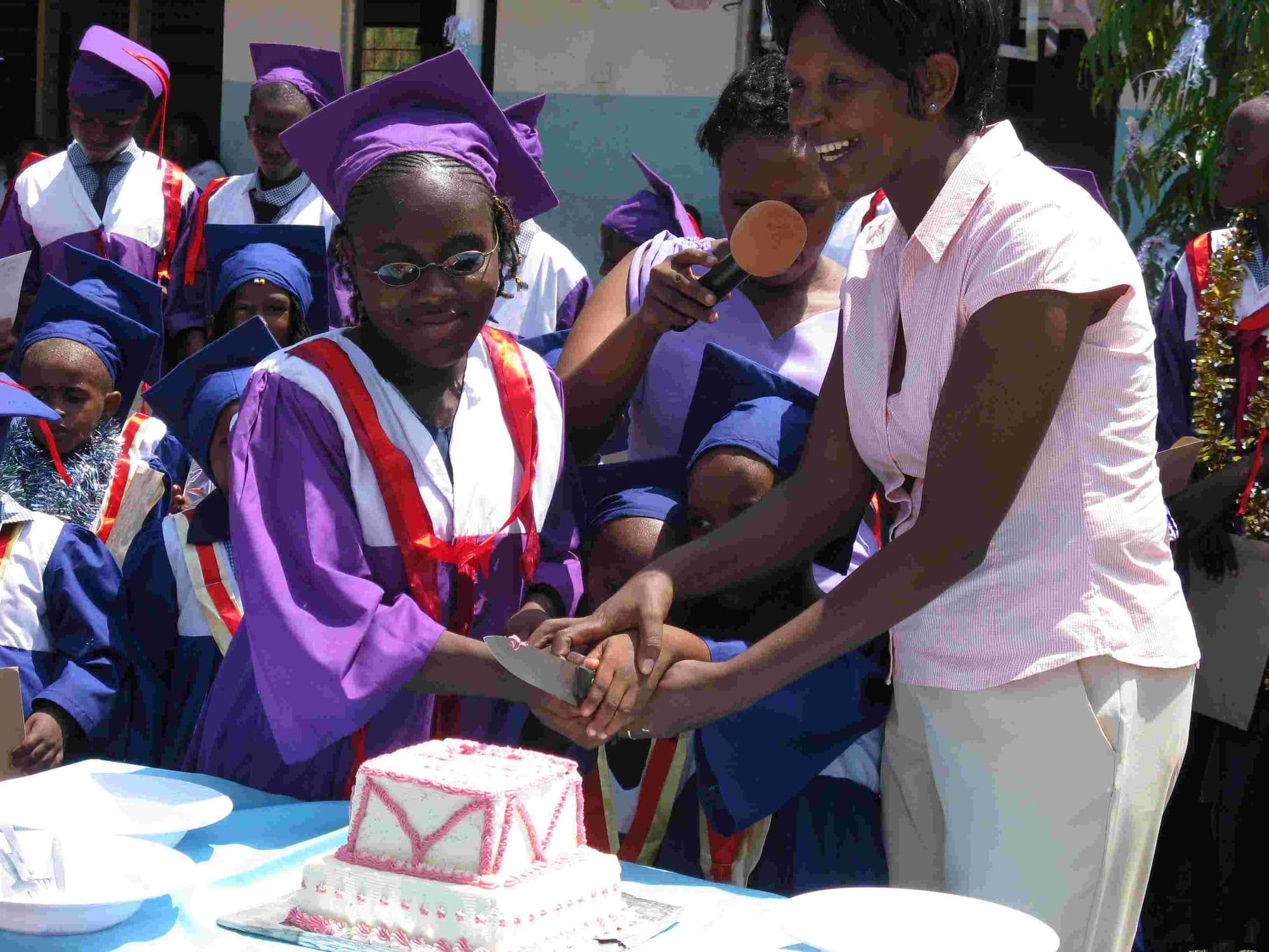 A graduate in a purple gown cutting a decorated cake with a teacher